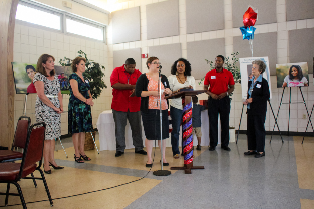 Eine Frau steht an einem Rednerpult mit einem Mikrofon, spricht, mit zwei leeren Stühlen daneben, einem Tisch mit weißer Decke, Topfpflanzen, einem roten und blauen, sternförmigen Ballon, Fotos auf Ständern und geschlossenen Fenstern im Hintergrund.