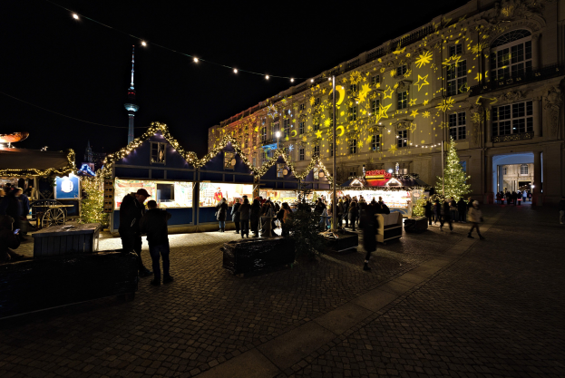 Ein geschäftiger Weihnachtsmarkt in Berlin, Deutschland, mit Menschen um dekorierte Stände und festliche Gebäude unter einem dunklen Himmel.