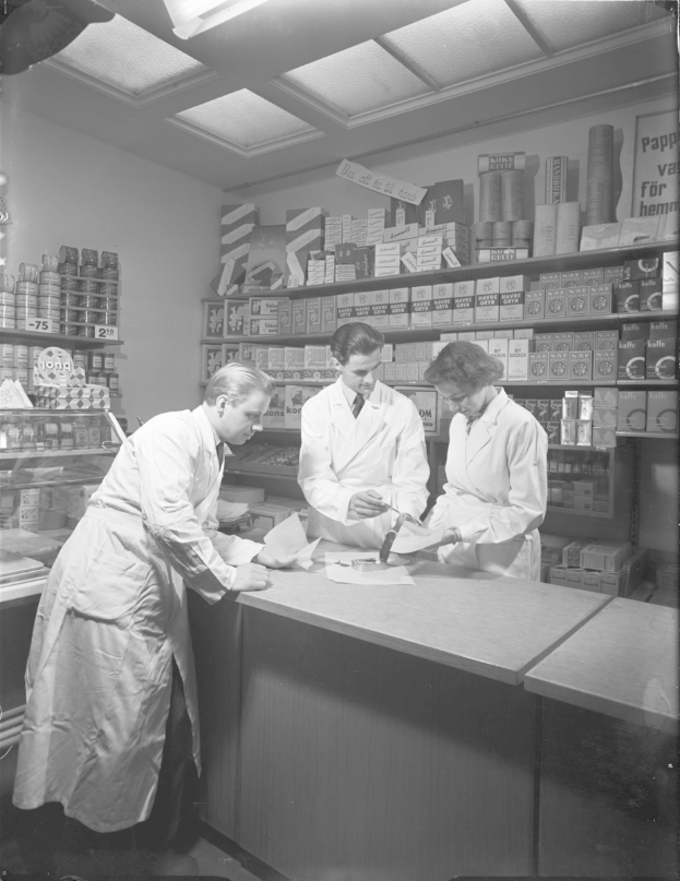 Drei Männer in weißen Kitteln an einer Apotheken-Theke mit Papieren und Gegenständen, vor Regalen mit Artikeln, in Schwarz-Weiß.