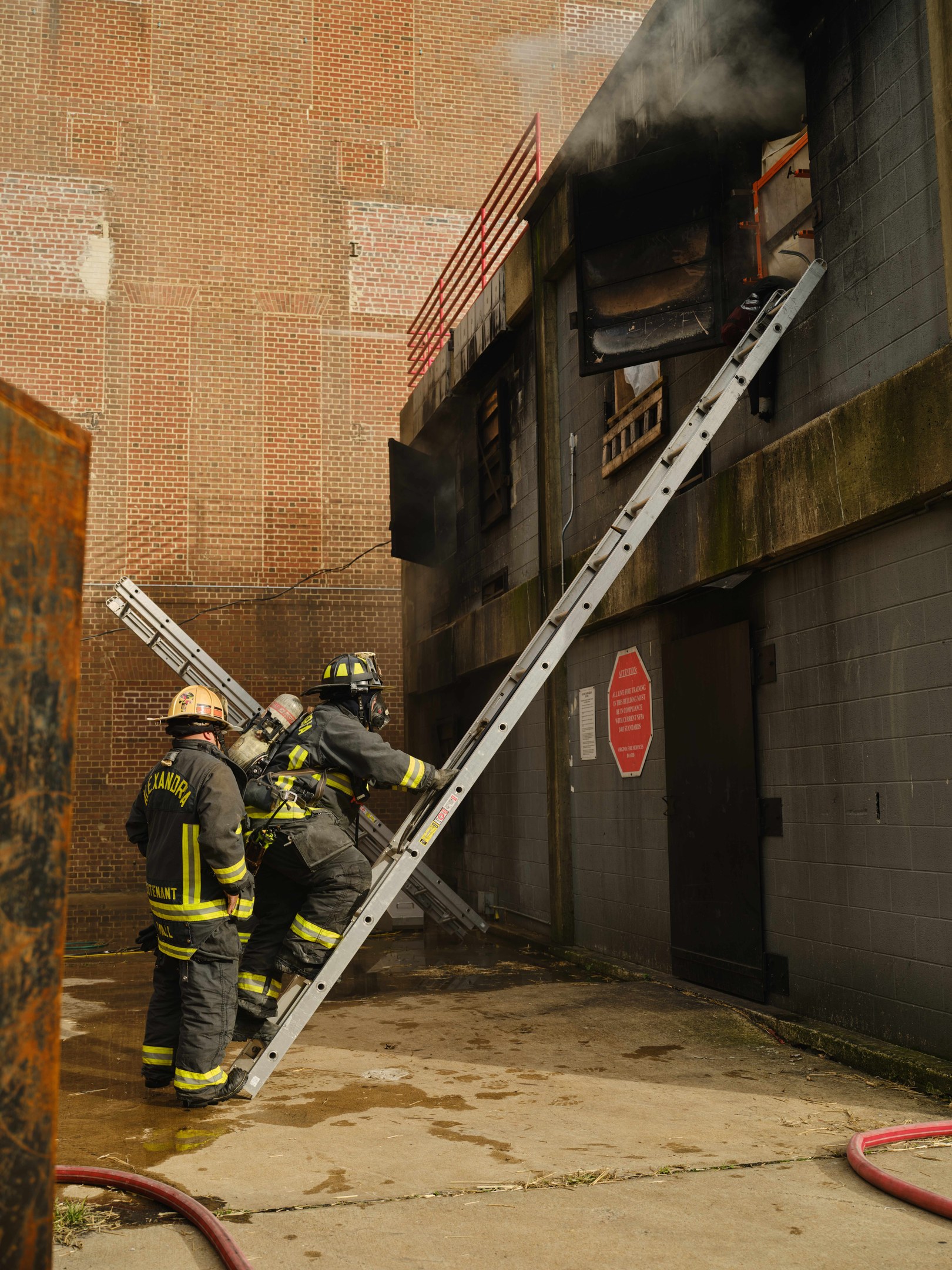 Feuerwehrleute in Helmen klettern eine Leiter zu einem rauchgefüllten Gebäude hinauf, mit einem Metallobjekt links und sichtbaren Rohren am Boden.