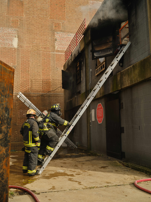 Feuerwehrleute in Helmen klettern eine Leiter zu einem rauchgefüllten Gebäude hinauf, mit einem Metallobjekt links und sichtbaren Rohren am Boden.