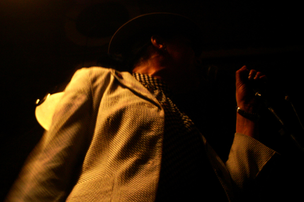 Ein Mann mit Hut und Jacke hält ein Mikrofon, das vor einem unscharfen Hintergrund hervorgehoben wird.