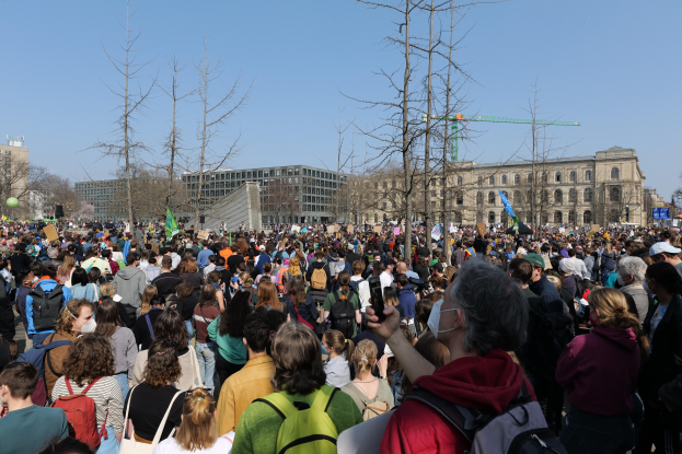 Eine große Menge von Menschen, die an einer Klimawandel-Demonstration vor einem Gebäude mit Fenstern, Bäumen und einem klaren Himmel teilnehmen, viele halten Schilder und tragen Taschen.