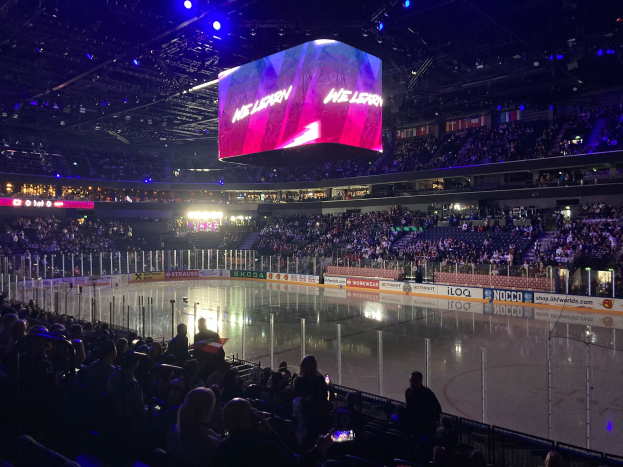 Ein Eishockeyspiel in einer großen Arena mit Zuschauern auf Sitzen und Stehplätzen, einem Zaun um die Eisfläche, einem Bildschirm mit Text und Deckenbeleuchtung.