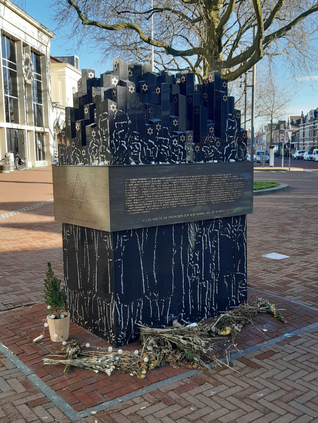 Denkmal in einem Stadtplatz für Holocaust-Opfer, mit einem Baum davor, einem Topfgewächs, verstreuten trockenen Blättern und Gebäuden, Fahrzeugen, Pfählen und Bäumen im Hintergrund.