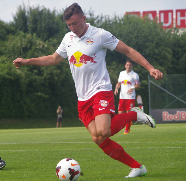 Ein professioneller Fußballspieler kickt einen Ball auf einem Rasenfeld mit Bäumen und einem klaren blauen Himmel im Hintergrund, mit einer Tafel mit der Aufschrift "RB Leipzig" im Bild.
