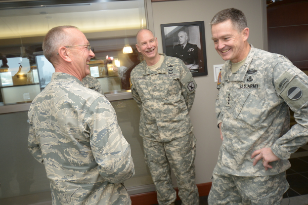 Drei uniformierte Männer stehen und lächeln vor einer Wand mit einem Foto Rahmen, einem Glasfenster, das Laternen und Tische zeigt, und einer braunen Tür auf der rechten Seite.