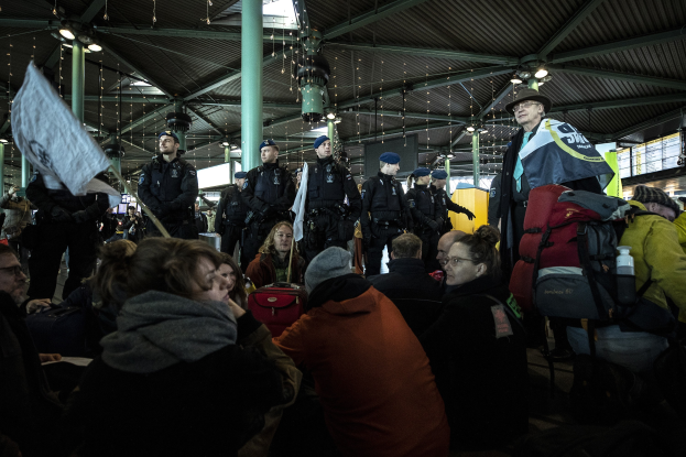 Eine Gruppe von Menschen steht vor einer Menge in einem Bahnhof, wobei einige Personen Schilder und Transparente halten, was auf eine Protestaktion hindeutet.