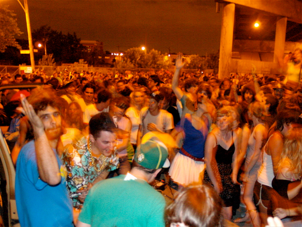 Menschen tanzen im Vordergrund eines Konzerts mit Lichtern, Pfählen, Bäumen und anderen Gegenständen im Hintergrund.