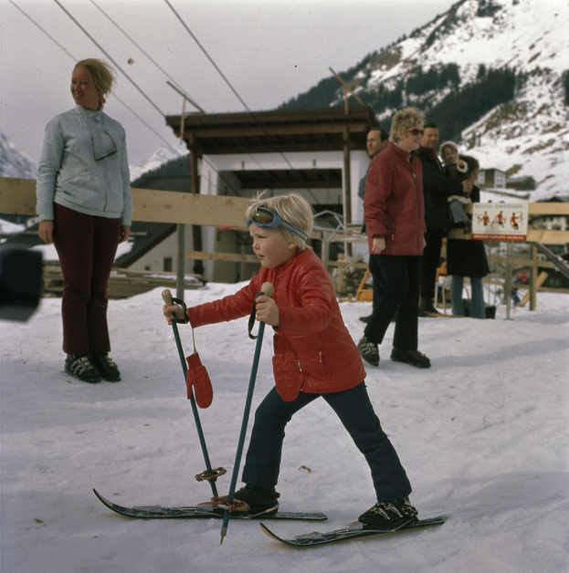 Ein junger Junge in einer roten Jacke fährt eine schneebedeckte Anhöhe mit Ski und Ski-Stöcken hinunter, mit Menschen, einem Holzzaun, einem Schuppen und einer Tafel mit Schrift im Hintergrund, unter einem sichtbaren Himmel.