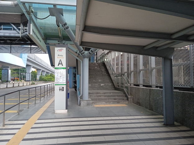 Ein Bahnsteig mit Treppe, eine Straße mit Geländern auf der linken Seite und eine Tafel mit Text oben auf dem Bahnsteig, mit Bäumen, Pfählen, Gebäuden und einer Brücke im Hintergrund.