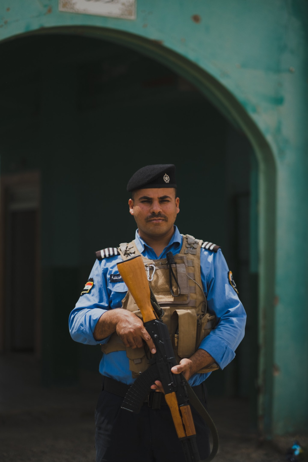 Polizeibeamter in Uniform mit Gewehr, vor einem Gebäude mit einem Tor und einer Tafel an der Wand.