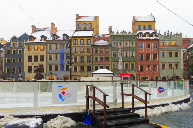 Eislaufbahn in einem Stadtplatz umgeben von Geb├Ąuden, Treppen, Gel├Ąndern, Informationsschildern, schneebedeckter Erde, Laternenmasten, B├Ąumen und einem Himmel als Hintergrund.