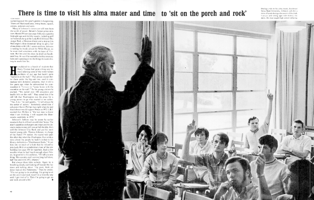 Eine Person, die steht und eine Brille trägt, mit vielen Menschen, die auf Stühlen in einem Raum mit Vorhängen und Fenstern sitzen.