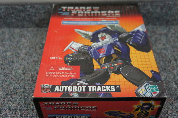 Ein roter, oranger und blauer Kasten mit einem Roboter und dem Text "AUTOBOT TRACKS" oben drauf, vor einem unscharfen schwarzen Hintergrund.