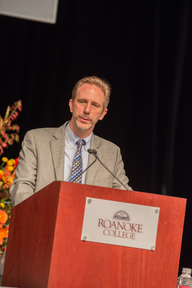 Ein Podium mit Mikrofonen und einer Flasche auf der rechten Seite, eine Person in der Mitte, Blumensträuße auf der linken Seite und ein Vorhang im Hintergrund.
