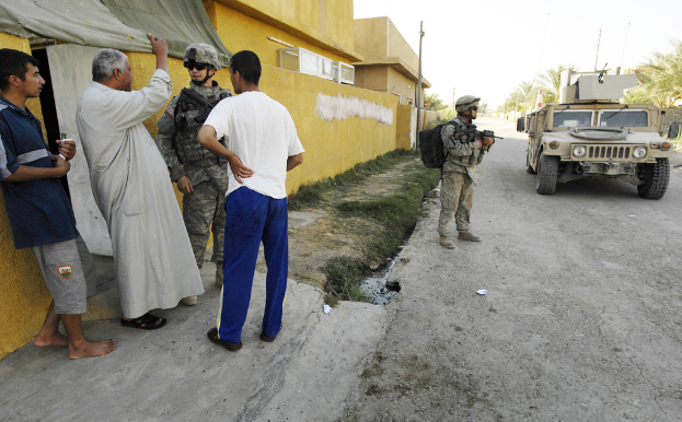 Eine Gruppe von Menschen steht im Freien, wobei eine Person eine Waffe hält, Häuser, Bäume, Pfosten, Wände und ein Fahrzeug im Hintergrund zu sehen sind.