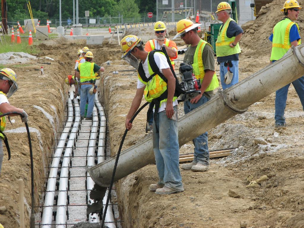 Eine Baustelle mit Arbeitern in Westen und Helmen, Absperrgittern, Zäunen, Bäumen, Pfählen, Erde und Rohren im Hintergrund.