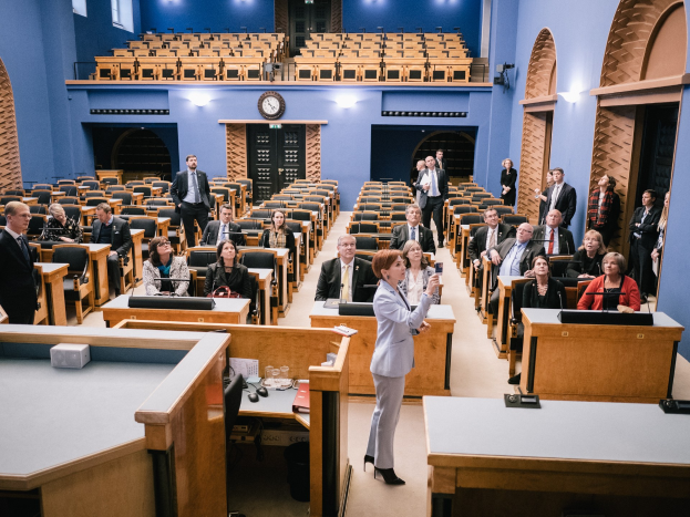 Frau hält Rede im schottischen Parlament, vor sitzenden und stehenden Zuhörern mit Tischen mit Mikrofonen, Gläsern, Papieren und anderen Gegenständen, unter Deckenlampen mit einer Uhr an der Wand.