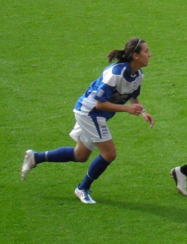 Eine Frau in einer blauen und weißen Uniform läuft auf einem Fußballfeld und trägt Fußballstiefel, wobei auf der rechten Seite des Bildes ein Personenschenkel zu sehen ist.