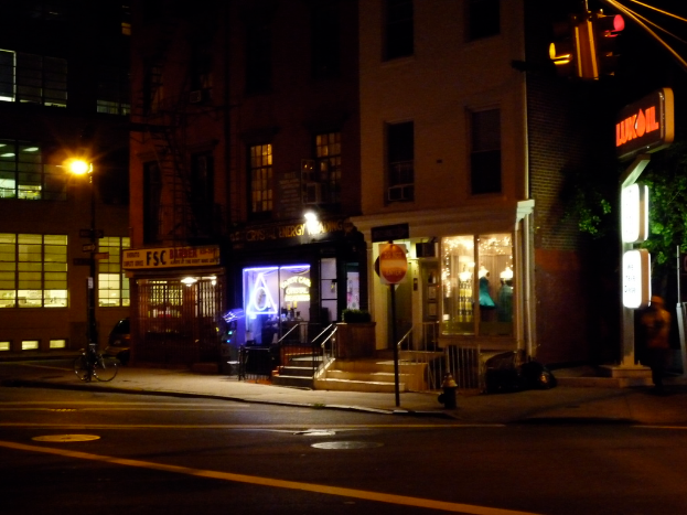 Eine Stadtansicht mit Gebäuden, beleuchteten Geschäften, Treppen, Bäumen, einer Straße, einem Fahrrad und einer Ampel.