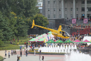 Ein gelber Helikopter fliegt ├╝ber einen Brunnen mit einer Menschenmenge, Zelten und Geb├Ąuden, B├Ąumen, Pflanzen, Gras und einem klaren blauen Himmel im Hintergrund.