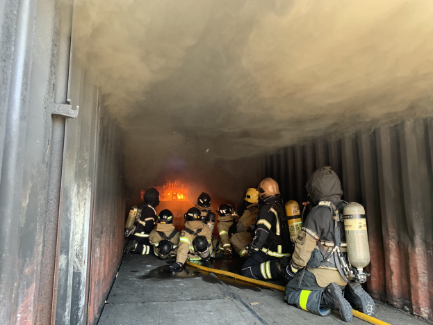 Feuerwehrleute in Schutzanzügen arbeiten in einem raucherfüllten Tunnel, einer hält einen Schlauch, mit sichtbaren Flammen im Hintergrund und einer Wand links.