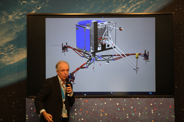 Ein Mann in einem weißen Hemd und schwarzer Blazer spricht in ein Mikrofon, vor einem Tisch und einer Projektionswand stehend, die einen robotischen Lander zeigt, mit einem blauen Himmel und Wolken im Hintergrund.