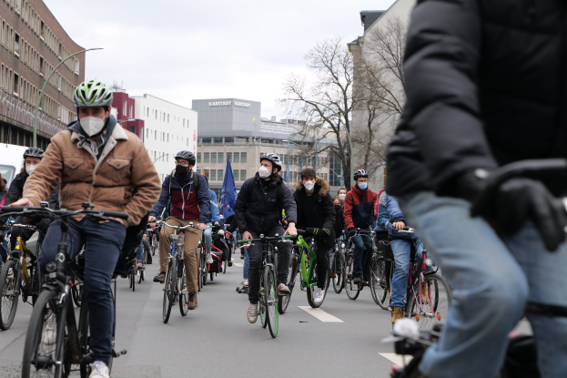 Eine Gruppe von Menschen in Helmen und Handschuhen fährt mit Fahrrädern eine von Bäumen gesäumte Straße in Berlin, Deutschland, entlang. Im Hintergrund sind Gebäude und ein geparktes Fahrzeug zu sehen.