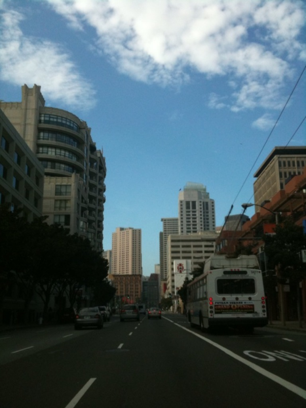 Eine belebte Straße mit verschiedenen Fahrzeugen, darunter ein Bus und Autos, Stromleitungen auf der rechten Seite, Gebäude im Hintergrund, Bäume auf der linken Seite und ein bewölkter Himmel oben.