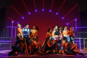 Eine Gruppe junger Tänzer in bunten Kostümen performs auf einer Bühne mit Geländern, beleuchtet von bunten Lichtern, während das Publikum jubelt und klatscht.