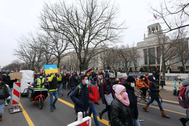 Eine große Gruppe von Menschen nimmt an einer Protestdemo in Washington, D.C. am 21. Januar 2020 teil und marschiert eine Straße entlang, wobei einige Schilder und Banner halten und andere Fahrräder fahren, mit Bäumen, Schildern und einem klaren blauen Himmel im Hintergrund.