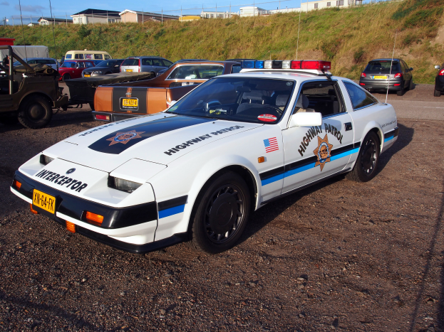 Ein Polizeiauto auf einem Parkplatz zwischen anderen Fahrzeugen geparkt, mit Gras, Pfählen, Drähten, Gebäuden und einem klaren blauen Himmel im Hintergrund.
