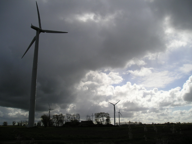 Windräder in einer Landschaft mit Bäumen, Wolken am Himmel und einem Haus in der Ferne.