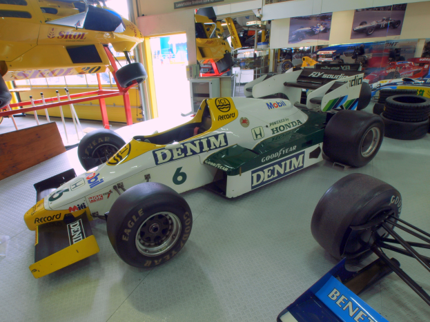 Eine Garage voller verschiedener Rennwagen, Reifen, ein hölzernes Objekt, ein Geländer, eine Glastür, eine Wand mit Fotorahmen und Deckenleuchten.