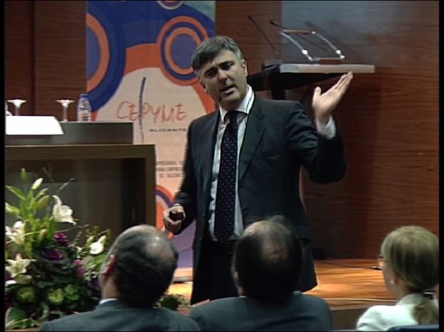 Ein Mann in einer schwarzen Jacke steht an einem Podium mit Mikrofonen, spricht in ein Handmikrofon, mit einer Blumenanordnung und einem Tisch mit einem Buch und einer Wasserflasche in der Nähe, während drei Personen auf Stühlen vor ihm sitzen, vor einem Hintergrund aus einer braunen Wand und einer bunten Tafel mit Text.