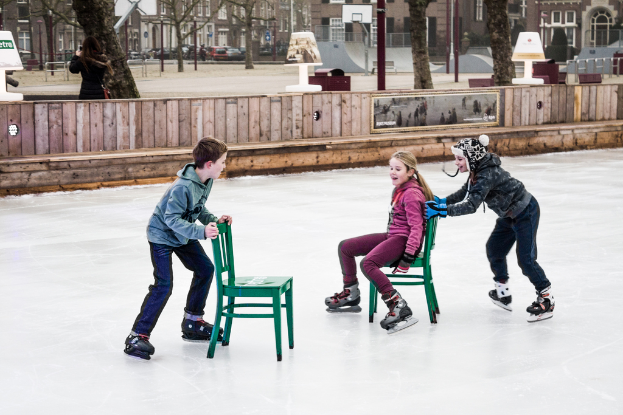 Kinder beim Skifahren vor einem Spielplatz im Hintergrund mit drei Kindern und zwei Stühlen in der Mitte und Gebäuden, Bäumen, Bänken, Pfosten und einem Basketballfeld im Hintergrund.