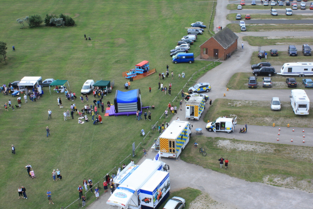 Eine Vogelperspektive eines Parkplatzes mit vielen Fahrzeugen, einigen Menschen in der Nähe von Zelten, einem zentralen Zelt und einigen Bäumen auf der linken Seite.