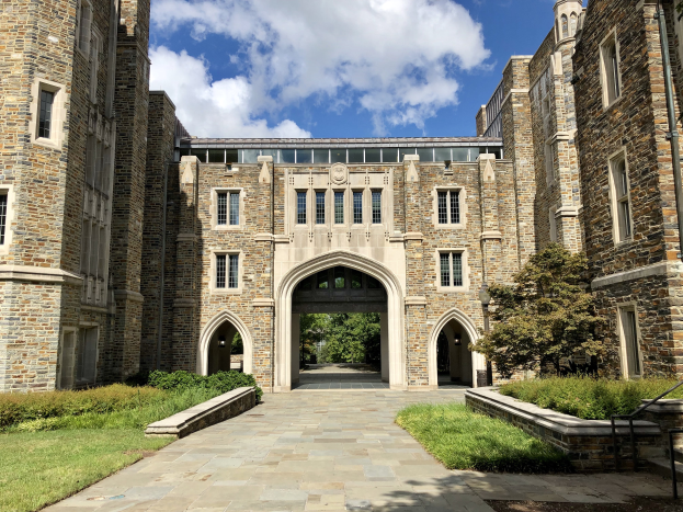Ein großer steinerner Universitätsgebäude mit einem Weg, Fenstern und einem Bogen, umgeben von Grün, mit einem bewölkten Himmel im Hintergrund.