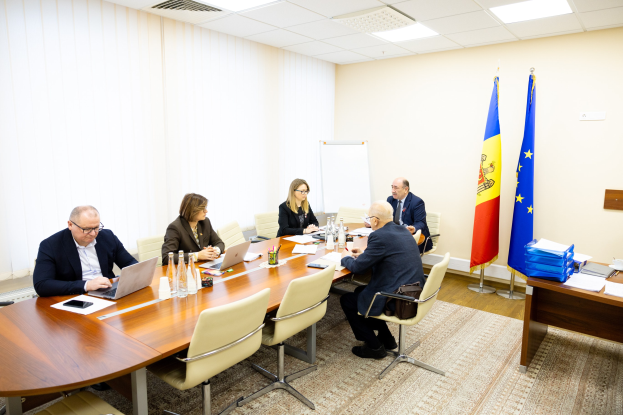 Eine Gruppe von Menschen sitzt um einen Konferenztisch mit Laptops, Flaschen, Gläsern, Papieren, Büchern und anderen Gegenständen und ist in eine Diskussion mit Flaggen, einer Whiteboard und Deckenbeleuchtung im Hintergrund vertieft.