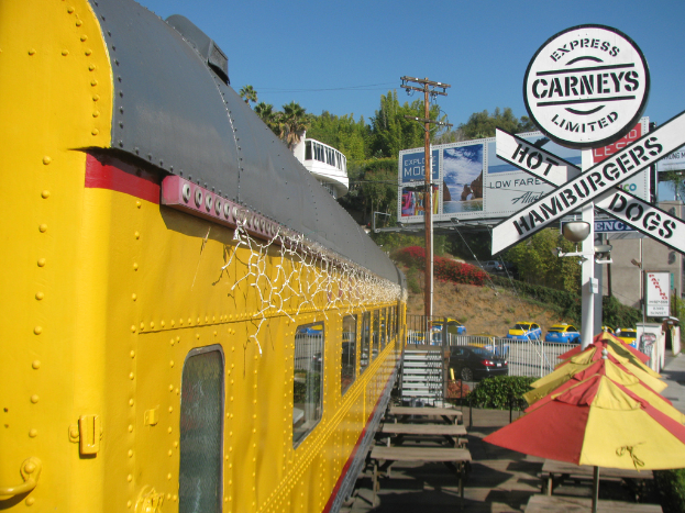 Eine mobile Kantine ist in der Nähe von Bänken und Stufen geparkt, mit Schirmen, Pflanzen und einem Geländer in der Nähe. Fahrzeuge, ein Pfahl, Schilder und Werbetafeln sind ebenfalls zu sehen, sowie ein Gebäude, Bäume und der Himmel im Hintergrund.