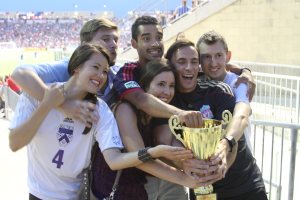 Sechs Personen, zwei Frauen und vier Männer, die eine Trophäe halten und lächeln, posieren für ein Foto vor einem Eisengeländer, mit einer Wand und einem Geländer oben rechts, Menschen und Lichter im Hintergrund, möglicherweise in einem Stadion.