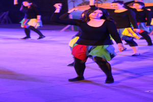 Eine Gruppe junger Frauen tanzt auf einer Bühne mit einem Gemälde an der Wand im Hintergrund, alle tragen Schuhe.