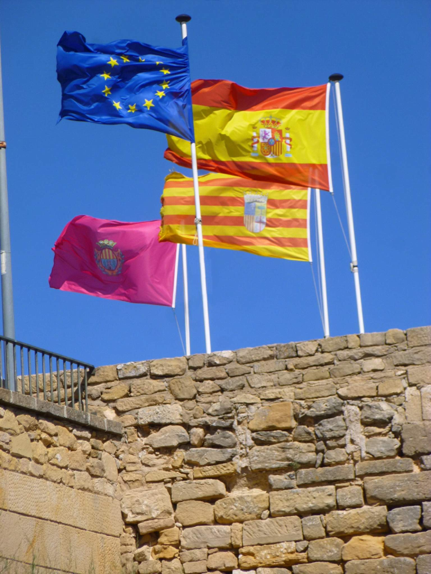 Drei Flaggen Spaniens und der Europäischen Union vor einer Steinmauer, mit einem Geländer auf der linken Seite und einem klaren blauen Himmel im Hintergrund.