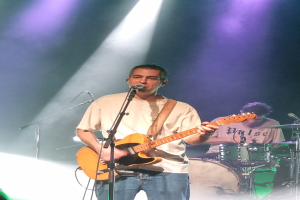 Ein Mann spielt eine E-Gitarre und singt in ein Mikrofon auf der Bühne, während ein anderer Mann Schlagzeug spielt.