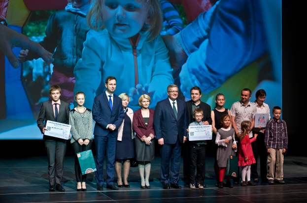 Eine Gruppe von Menschen auf einer Bühne, einige halten Taschen und Urkunden, mit einem Bildschirm im Hintergrund, der ein Bild eines Kindes und anderer Menschen anzeigt, wahrscheinlich bei einem Charity-Event.