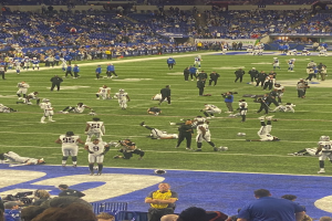 Ein Fußballspiel in einem Stadion mit Menschen auf dem Feld und einigen im Hintergrund sitzend, mit einer Person, die eine Kamera hält, was eine Live-Übertragung anzeigt.