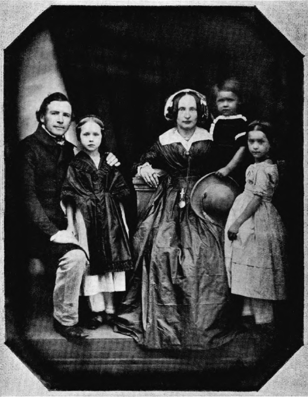 Ein Schwarz-Weiß-Foto einer Familie, die auf einer Treppe posiert, mit einer Frau in einem Kleid, die einen Hut hält und zwei Kinder neben ihr stehen.