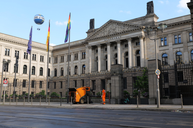Großes modernes Gebäude mit vielen Fenstern, als Bundestag in Berlin identifiziert, umgeben von Straßeninfrastruktur, Fußgängern und Flaggen, mit Bäumen und einem bewölkten Himmel im Hintergrund.