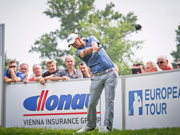 Ein Mann mit Mütze schwingt einen Golfschläger auf einem grünen Feld, mit Text- und Logo-Tafeln, Bäumen und einem klaren blauen Himmel im Hintergrund, was das Finale der European Tour anzeigt.